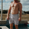 Man wearing gray swim shorts standing by a pool with a palm tree and house in the background.
