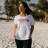 Woman wearing a white t-shirt with 'Swearing Helps' text on a sandy beach.
