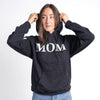 Person wearing a black hoodie with 'MOM' printed on it against a white background