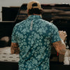 Man wearing a teal floral shirt with a visible brand logo, standing in front of a vehicle.