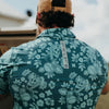 Person wearing a teal floral patterned shirt with a brand label outdoors.