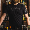 Person wearing a black t-shirt with 'Whiskey Helps' text in a bar setting.