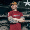 Man wearing a red t-shirt with 'American' text and a cap, standing in front of a military vehicle.