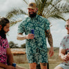 Man in a floral shirt holding a can, standing between two others outdoors with palm trees in the background