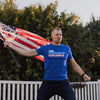 Man holding an American flag and wearing a blue t-shirt with 'The Goat' print outdoors.