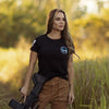 Woman holding a rifle in a natural setting with greenery