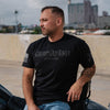 A man wearing a black graphic t-shirt with 'Grumpy Old Biker' printed on the front, sitting outdoors next to a vehicle with a concrete wall and city buildings in the background.