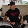A man wearing a black t-shirt with red 'Strokes COUNT' text and a black cap sits on a white motorcycle in an outdoor setting.
