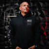 A man wearing a black pullover hoodie with a white American flag and 'Guns & Ammo' logo on the left chest, and 'THIS WILL DEFEAT' text on the right sleeve, posed against a dark geometric wall.