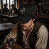 Man in a cap sitting at a bar holding a mug, with a rustic interior.