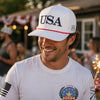 Man wearing a white cap with 'USA' and a white t-shirt with a logo, smiling outdoors.