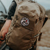 Brown backpack with a patch on a person's back, outdoors.