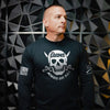 A man wearing a black long-sleeve t-shirt with a white skull and crossbones graphic on the chest that reads 'THE GOONS ALWAYS SAY KILL'. The shirt also features a US flag patch on the right sleeve and a tactical patch on the left sleeve, set against a dark geometric wall.