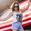 Woman holding an American flag with a graphic tank top and denim shorts.
