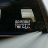 A white vinyl decal with the text 'SOMEONE STILL ANSWERS THE CALL' and a small blue and red accent bar, applied to the rear window of a vehicle.