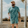 Man wearing a teal floral shirt and shorts set with a cap, standing outdoors.