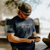 Man holding a handgun outdoors, wearing a t-shirt with text and an American flag design.
