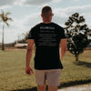A man viewed from behind wearing a black t-shirt with white printed text on the back and light beige shorts, standing outdoors in a grassy field under a cloudy sky.