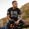 Man wearing a black t-shirt with 'Girl Dad' text, sitting outdoors with a child and rifle.