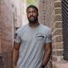 Man wearing a gray t-shirt with a logo, standing in a narrow alleyway.