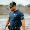 Man wearing a navy blue t-shirt with a logo and text, standing outdoors near water.