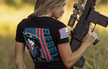 Person wearing a black t-shirt with a graphic design and text, holding a rifle in an outdoor setting.
