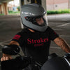 A man wearing a gray full-face motorcycle helmet and a black t-shirt with red 'Strokes COUNT' text sits on a motorcycle in a parking structure.