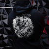 A person shown from the back wearing a black hoodie with a large distressed white graphic on the back featuring a skull, barbed wire, and a rifle. The background is a dark geometric wall with red accents.