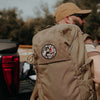 Person wearing a tan backpack with a logo, standing outdoors near a vehicle.