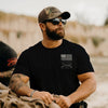 Man wearing a black t-shirt with a logo, sunglasses, and a cap in an outdoor setting.