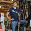 Man in a blue t-shirt and jeans standing next to a piece of industrial equipment in a warehouse.