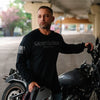 A man wearing a black long-sleeve t-shirt with 'Grumpy Old Biker' printed on the chest stands next to a motorcycle in an outdoor underpass setting.