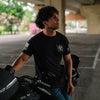 A man wearing a black short-sleeve t-shirt with a small skull graphic on the left chest and a striped patch on the right sleeve, sitting on a black motorcycle in a parking structure.