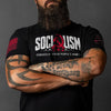 A bearded man with tattooed arms wearing a black graphic t-shirt. The shirt features the word 'SOCIALISM' in white block letters with a red ribbon symbol integrated into the lettering, and the tagline 'POWERED BY OTHER PEOPLE'S MONEY' underneath.