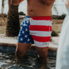 Person wearing American flag swim shorts standing in water with palm trees in the background