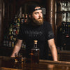 Man in a bar wearing a 'Whiskey Helps' shirt with a bottle and glasses on the counter.