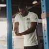 Man in a gym setting wearing a white t-shirt with a logo, interacting with a white bucket.