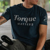 A man wearing a dark navy blue heather t-shirt with 'Torque MATTERS' printed in white across the chest, seated on a motorcycle.