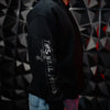 A person wearing a black hoodie with a distressed white graphic on the left sleeve that reads 'THIS WILL FELL' with a frog illustration, standing in front of a dark geometric wall.