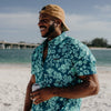 Man wearing a blue floral shirt and beige bucket hat on a beach.