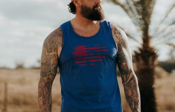 Man wearing a blue tank top with an American flag design, standing outdoors with palm trees in the background.