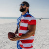 Man wearing a red, white, and blue shirt holding a football on a beach.