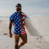 Man wearing a USA flag shirt and sunglasses, holding a surfboard on a beach.