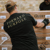 Person wearing a black t-shirt with 'Forward with Purpose' on the back, working with stacked boxes.