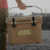Person holding a beige cooler with 'Over Taxed' label outdoors