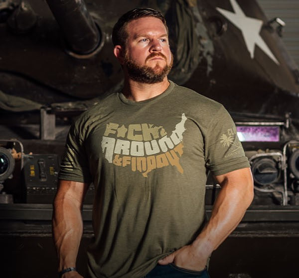 A muscular man with a beard wearing an olive green graphic t-shirt that reads 'F*CK AROUND & F*DD IT' in a distressed stencil font, standing in front of a dark military tank.