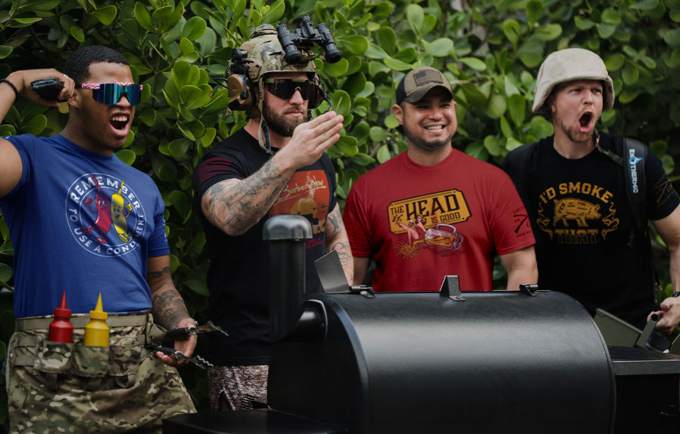 Four men in tactical gear and casual clothing standing around a barbecue grill with a green leafy background.