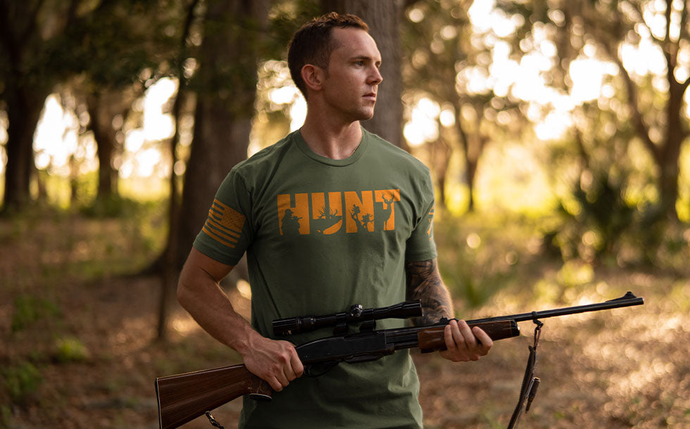 A man in a forest setting wearing an olive green t-shirt with a distressed orange 'HUNT' graphic on the chest, holding a rifle with a scope.