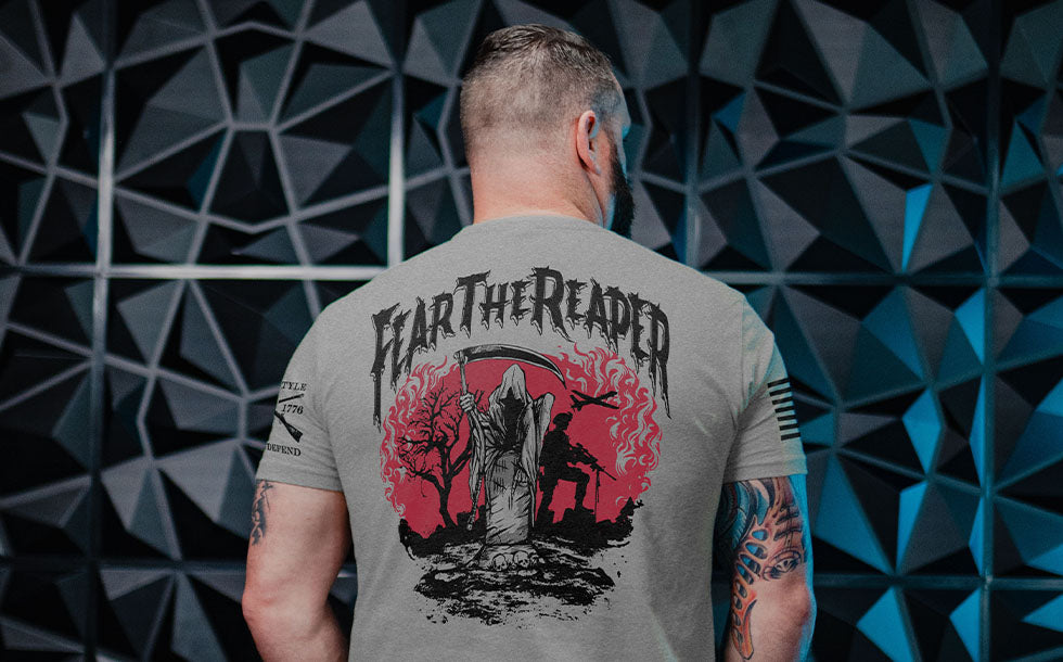 A man viewed from behind wearing a heather gray t-shirt with a large back graphic that reads 'HEAR THE READER' above a grim reaper and tombstone design in black and red. He is standing in front of a dark geometric wall.