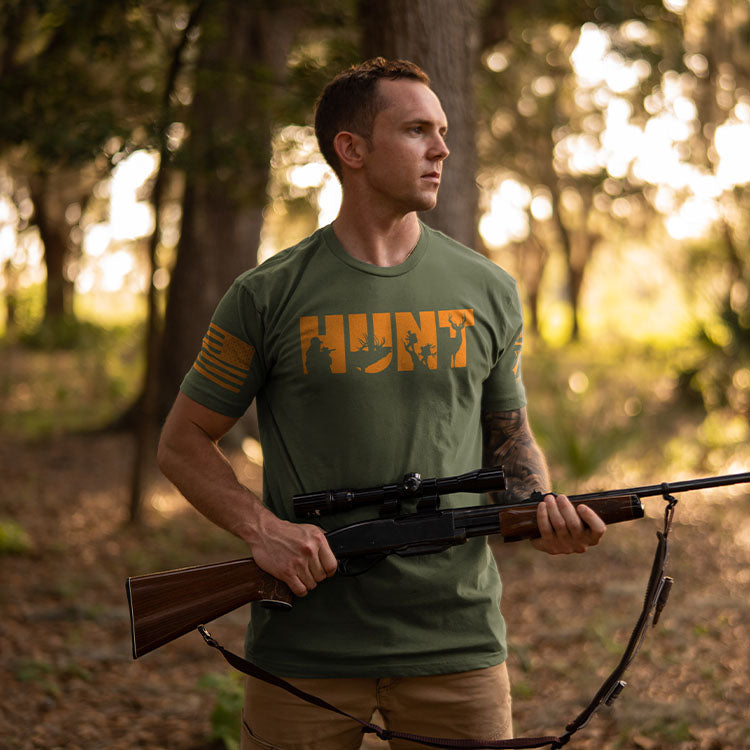 A man in a forest setting wearing an olive green t-shirt with a distressed orange 'HUNT' graphic across the chest, holding a rifle with a scope.